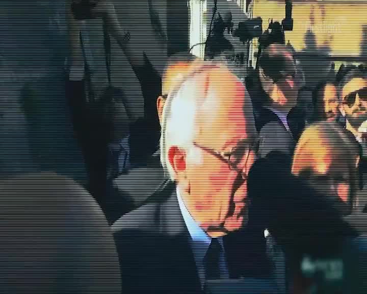 The morning sun catches the glint of glasses on a man in a suit, his head turned as he moves through a crowd. Cameras are held high, their lenses pointed forward, ready to capture the moment.