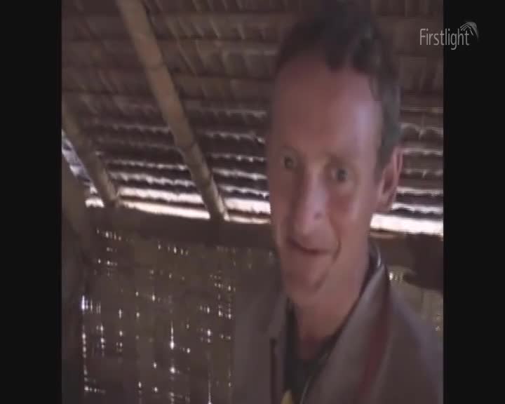 Sunlight streams through the woven walls of a rustic shelter. A man with a friendly smile looks directly at me.