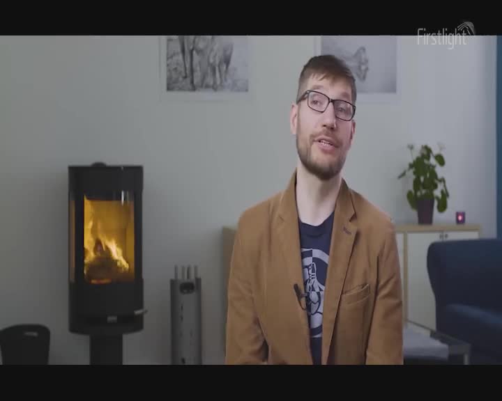 Flames dance in the glass-fronted wood burner. A man in a tan jacket speaks, his glasses reflecting the light.