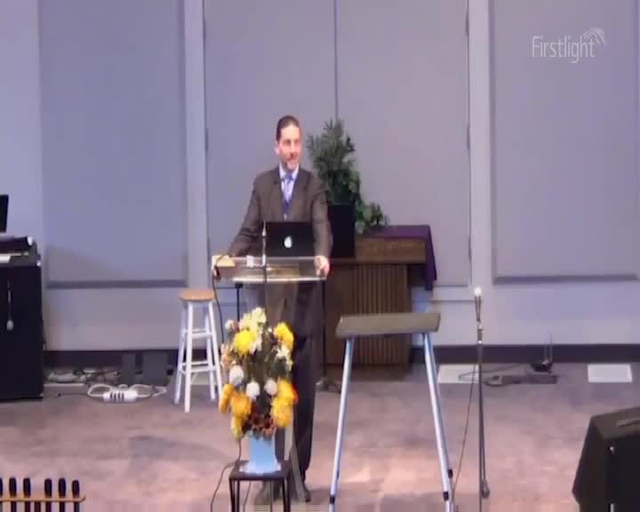 A man in a suit stands behind a lectern, speaking into a microphone. A laptop sits open on the lectern, and a vase of bright yellow and orange flowers is on a small table to his left.