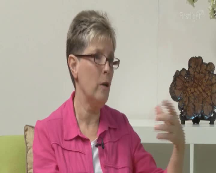 A woman with short, grey hair and glasses speaks animatedly, her right hand gesturing outwards. Behind her, a decorative piece resembling a dark, textured fern rests on a shelf.
