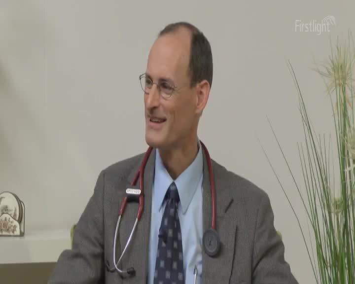 A man in a suit and tie, wearing glasses and a stethoscope, speaks with a slight smile. Tall, feathery grass stands to his right. A man in a suit and tie, wearing glasses and a stethoscope, speaks with a slight smile. Tall, feathery grass stands to his right.