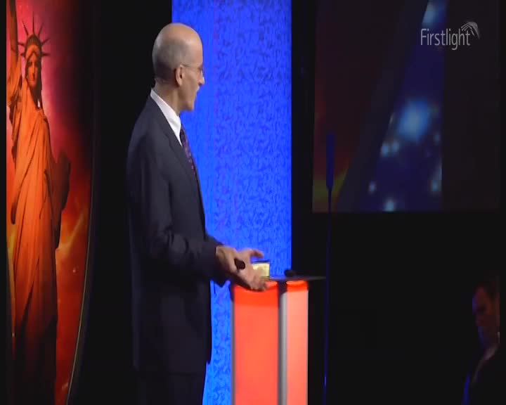 A man in a dark suit stands on a stage, holding a small yellow object. Behind him, a large image of the Statue of Liberty glows with fiery orange light. A man in a dark suit stands on a stage, holding a small yellow object. Behind him, a large image of the Statue of Liberty glows with fiery orange light.
