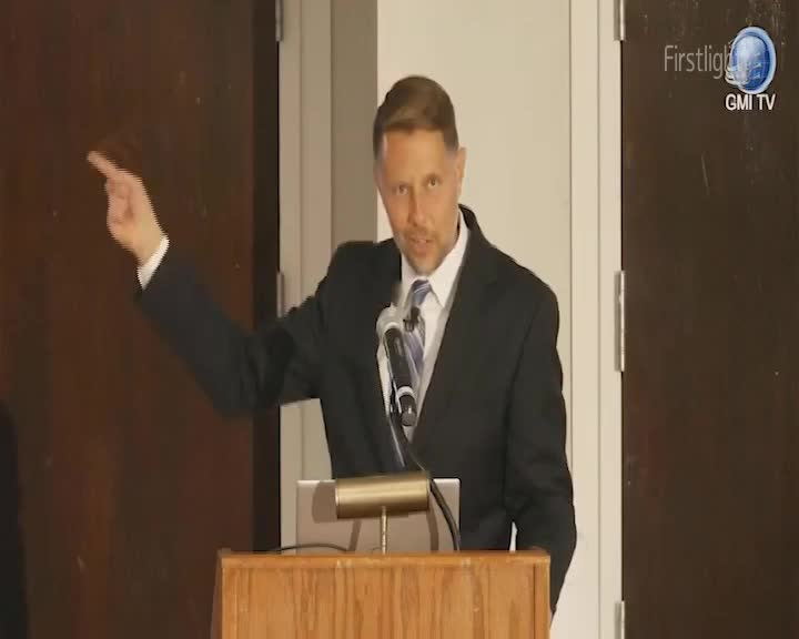 A man in a suit gestures with his left hand, pointing towards something off-camera. He stands behind a wooden lectern, a microphone positioned before him. A man in a suit gestures with his left hand, pointing towards something off-camera. He stands behind a wooden lectern, a microphone positioned before him.