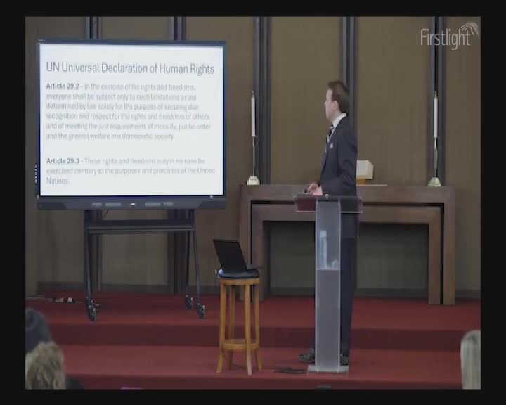 A man in a suit stands at a podium, gesturing towards a large screen displaying the UN Universal Declaration of Human Rights. The room is lit by soft, early morning light, and the Firstlight logo is visible in the corner. A man in a suit stands at a podium, gesturing towards a large screen displaying the UN Universal Declaration of Human Rights. The room is lit by soft, early morning light, and the Firstlight logo is visible in the corner.