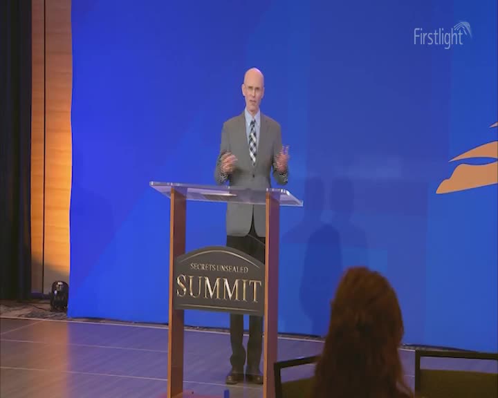 A bald man in a grey suit gestures with his hands as he speaks from behind a podium. The "Secrets Unsealed Summit" is emblazoned on the front of the wooden stand. A bald man in a grey suit gestures with his hands as he speaks from behind a podium. The "Secrets Unsealed Summit" is emblazoned on the front of the wooden stand.