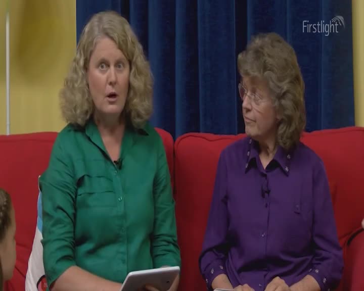Two women are sitting on a red couch, bathed in the soft light of early morning. The woman on the left, wearing a green shirt, holds a tablet, her mouth open as if speaking. Beside her, a woman in a purple shirt looks off to the side, her glasses perched on her nose. Two women are sitting on a red couch, bathed in the soft light of early morning. The woman on the left, wearing a green shirt, holds a tablet, her mouth open as if speaking. Beside her, a woman in a purple shirt looks off to the side, her glasses perched on her nose.