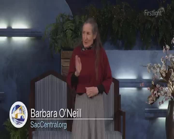 A woman stands on a stage, her hand raised as if making a point. Behind her, lush ferns and a vase of delicate flowers frame the scene. A woman stands on a stage, her hand raised as if making a point. Behind her, lush ferns and a vase of delicate flowers frame the scene.