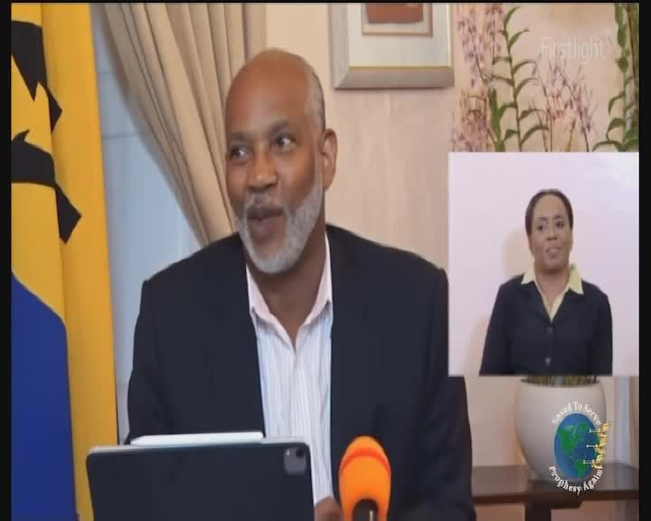 A man with a grey beard speaks into a microphone, the Barbados flag behind him. A smaller screen shows a woman signing to him. A man with a grey beard speaks into a microphone, the Barbados flag behind him. A smaller screen shows a woman signing to him.