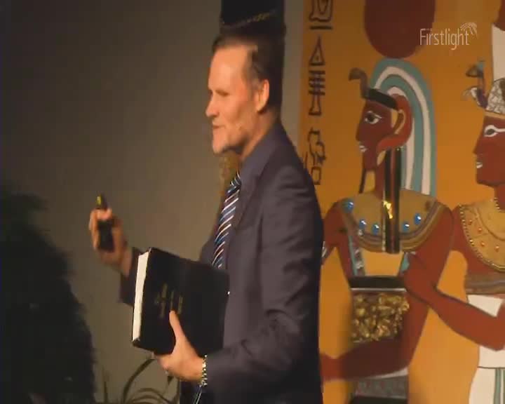 A man in a suit stands holding a thick book and a clicker. Behind him, a mural depicts figures with Egyptian headdresses. A man in a suit stands holding a thick book and a clicker. Behind him, a mural depicts figures with Egyptian headdresses.
