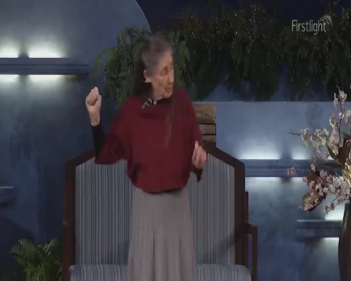 A woman in a burgundy top and grey skirt gestures with her hands. She stands on a stage with a backdrop of blue panels and greenery. A woman in a burgundy top and grey skirt gestures with her hands. She stands on a stage with a backdrop of blue panels and greenery.