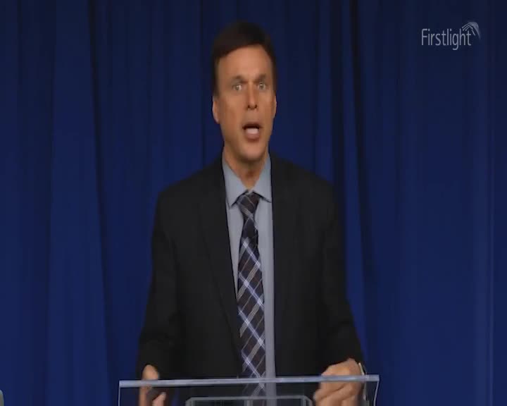 A man in a suit speaks animatedly behind a lectern. The blue curtain behind him is subtly lit.