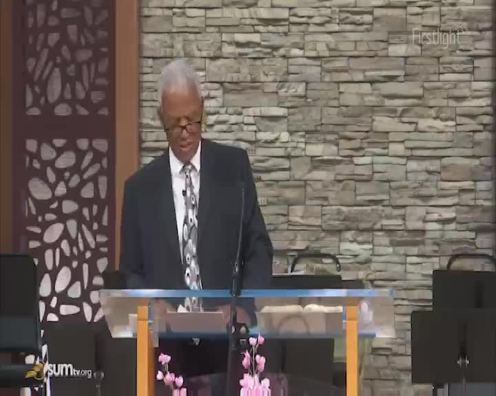 A man in a suit stands at a podium, his gaze fixed on something ahead. The stone wall behind him is illuminated by the early morning light.