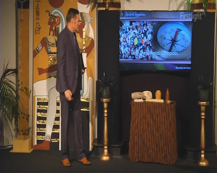 A man in a dark suit walks across the stage, his right hand holding a white folder. Behind him, a large screen displays a compass and the words "Lost Their Moral Compass."