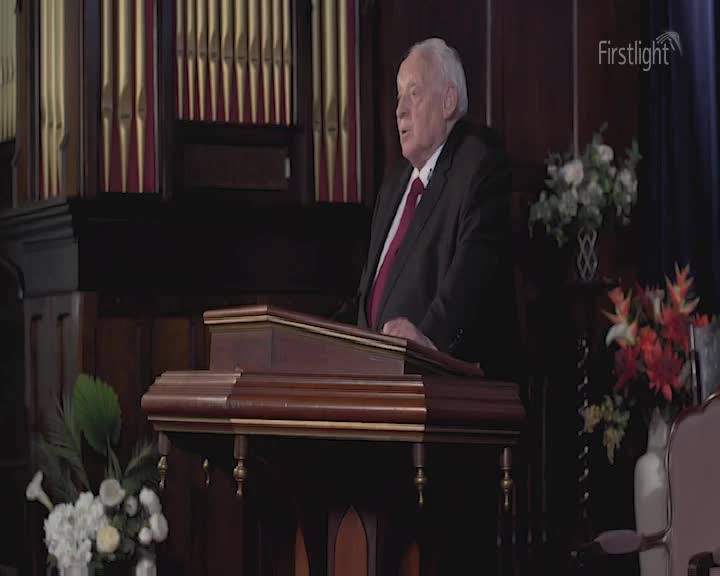 An older man in a suit and tie stands at a wooden lectern, speaking. Behind him, a pipe organ fills the space.