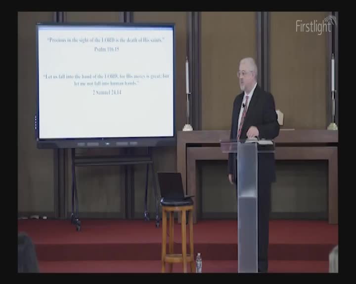 A man in a suit stands at a podium, his hand resting on a Bible. A large screen behind him displays scripture.