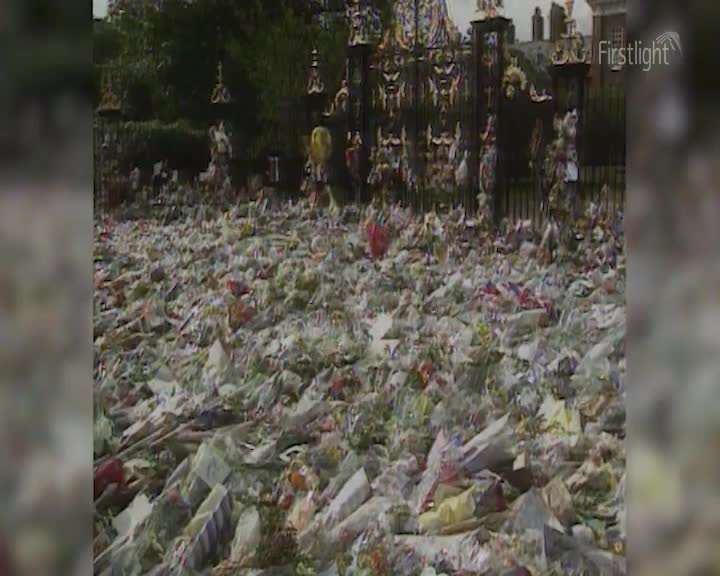 The ground is covered in a thick carpet of floral tributes, a sea of white and pastel blooms stretching towards an ornate black gate.  The air is heavy with the scent of a thousand bouquets, a silent testament to a moment of shared grief.