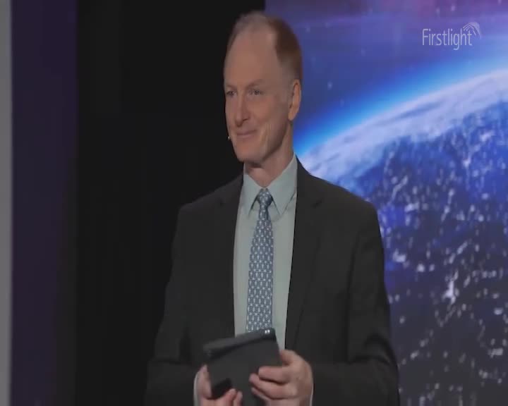 A man in a suit stands on a stage, holding a tablet. Behind him, a large screen displays a view of Earth from space, with the "Firstlight" logo in the corner.