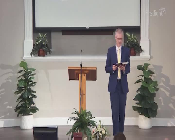 A man in a suit stands at a wooden lectern, holding a book. Two large potted plants flank the stage, their leaves reaching towards the ceiling.