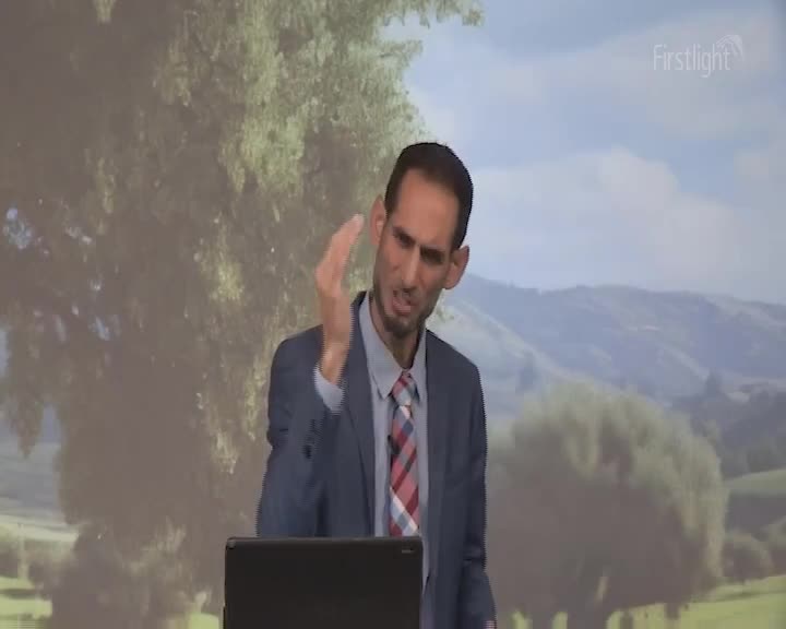 A man in a suit gestures emphatically, his hand moving through the air. Behind him, rolling green hills under a soft sky evoke a New Zealand landscape.