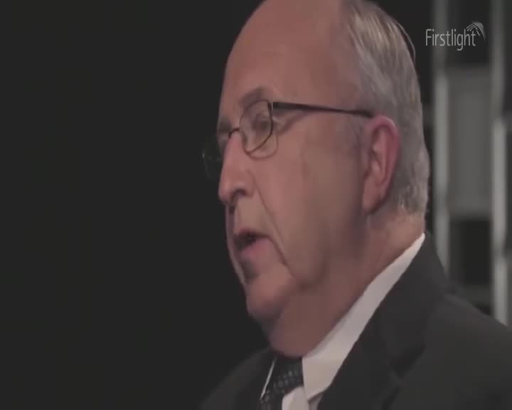 A man in a dark suit and tie speaks, his glasses reflecting the light. The Firstlight logo appears in the upper right corner.