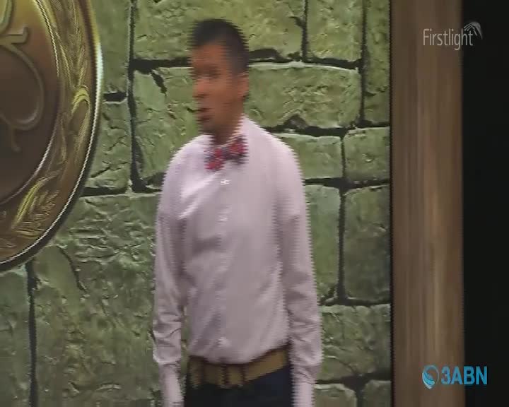 A man in a light pink shirt and a red bow tie stands before a stone wall. A large, golden medallion is visible to his left.