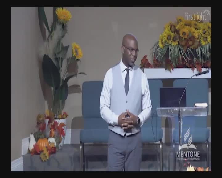 A man stands at a podium, hands clasped, with sunflowers and autumnal decorations behind him. The light from the Firstlight broadcast illuminates the scene.