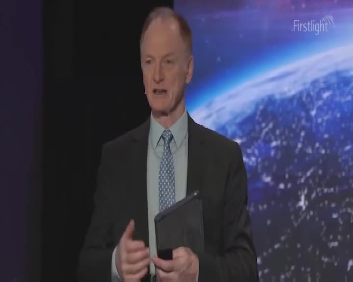 A man in a suit stands speaking, gesturing with his hands. Behind him, a projection displays a view of Earth from space.