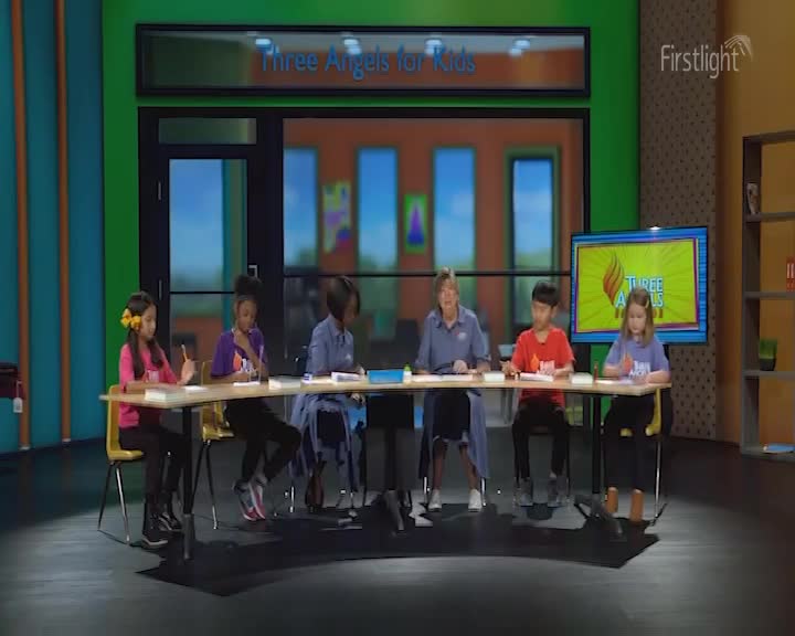 The children are seated at a table, each with a piece of paper and a pen. A woman in a blue shirt stands behind them, looking towards the camera. The Firstlight logo is visible in the upper right corner.