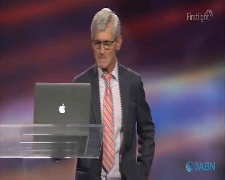 A man in a suit and tie stands at a podium with a laptop. The backdrop behind him glows with soft, diffused light.