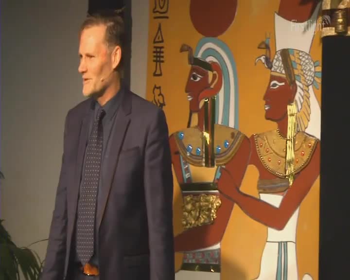 A man in a dark suit stands to the left of a vibrant mural depicting ancient Egyptian figures. The mural's warm, golden background is punctuated by the figures' rich red skin and elaborate headdresses.