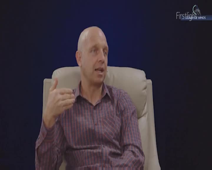 A bald man in a striped shirt gestures with his left hand as he speaks. He sits in a cream-colored chair against a dark blue background.