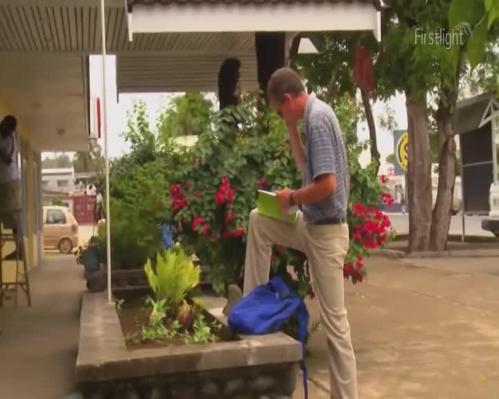 A man in a checkered shirt and tan trousers pauses by a flowerbed, holding a green notebook. He seems to be talking on his phone, his other hand resting on the notebook. A man in a checkered shirt and tan trousers pauses by a flowerbed, holding a green notebook. He seems to be talking on his phone, his other hand resting on the notebook.