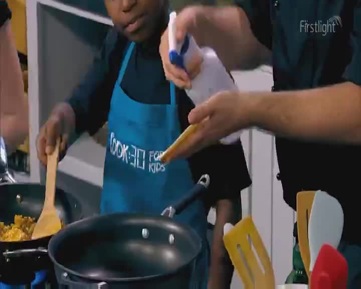 A young boy in a blue apron stirs food in a wok, while an adult sprays something onto a spatula. The Firstlight logo is visible in the upper right corner. A young boy in a blue apron stirs food in a wok, while an adult sprays something onto a spatula. The Firstlight logo is visible in the upper right corner.