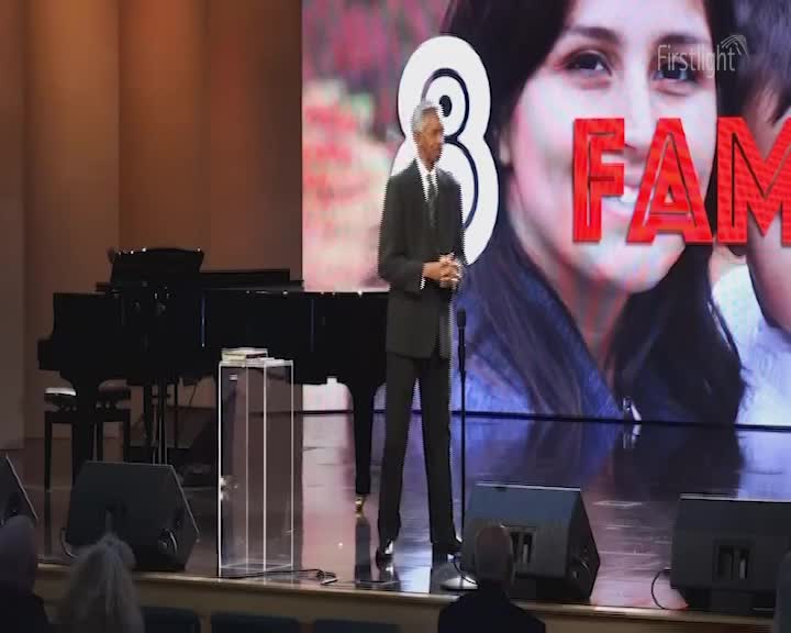 A man in a suit stands on a stage, speaking. Behind him, a large screen displays the word "FAMILY" over a photo of a woman.