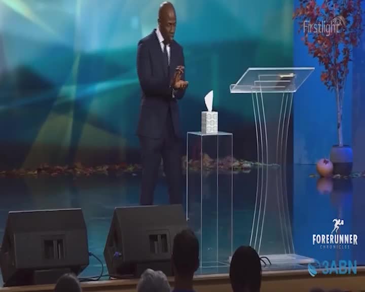 A man in a suit stands on a stage, holding a tissue. A clear podium and a box of tissues sit beside him.