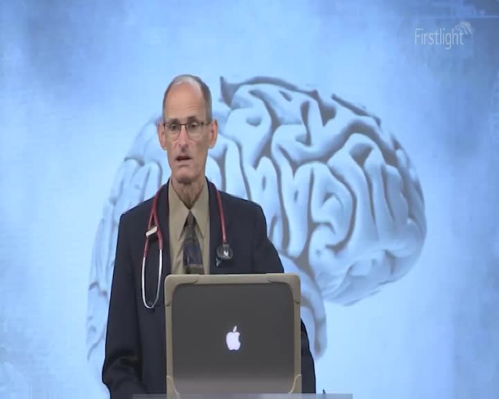 A man in a suit and tie, a stethoscope draped around his neck, stands before a large projection of a human brain. The Firstlight logo is visible in the upper right corner.
