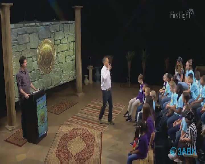 A man in a white shirt gestures as he speaks to a group of children seated on benches. Another man stands at a podium, facing the audience. The backdrop suggests a set designed to resemble an ancient stone wall.