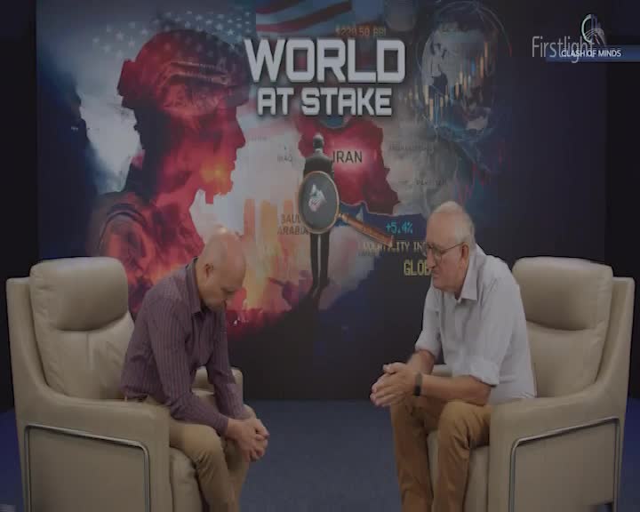 Two men sit in plush armchairs, facing each other. Behind them, a large screen displays a dramatic graphic with the words "WORLD AT STAKE."