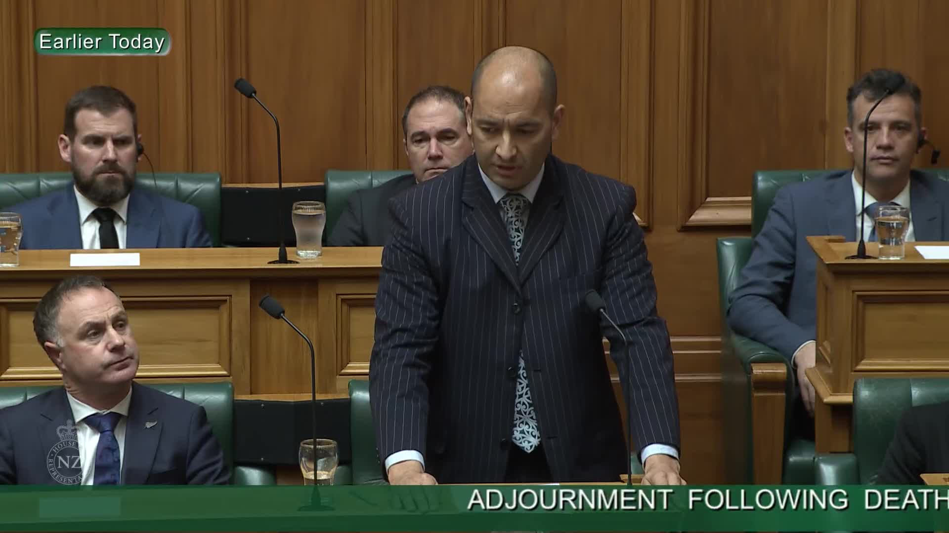 A man in a pinstripe suit stands at a podium, speaking into a microphone. Other men in suits sit behind him, some looking on intently.
