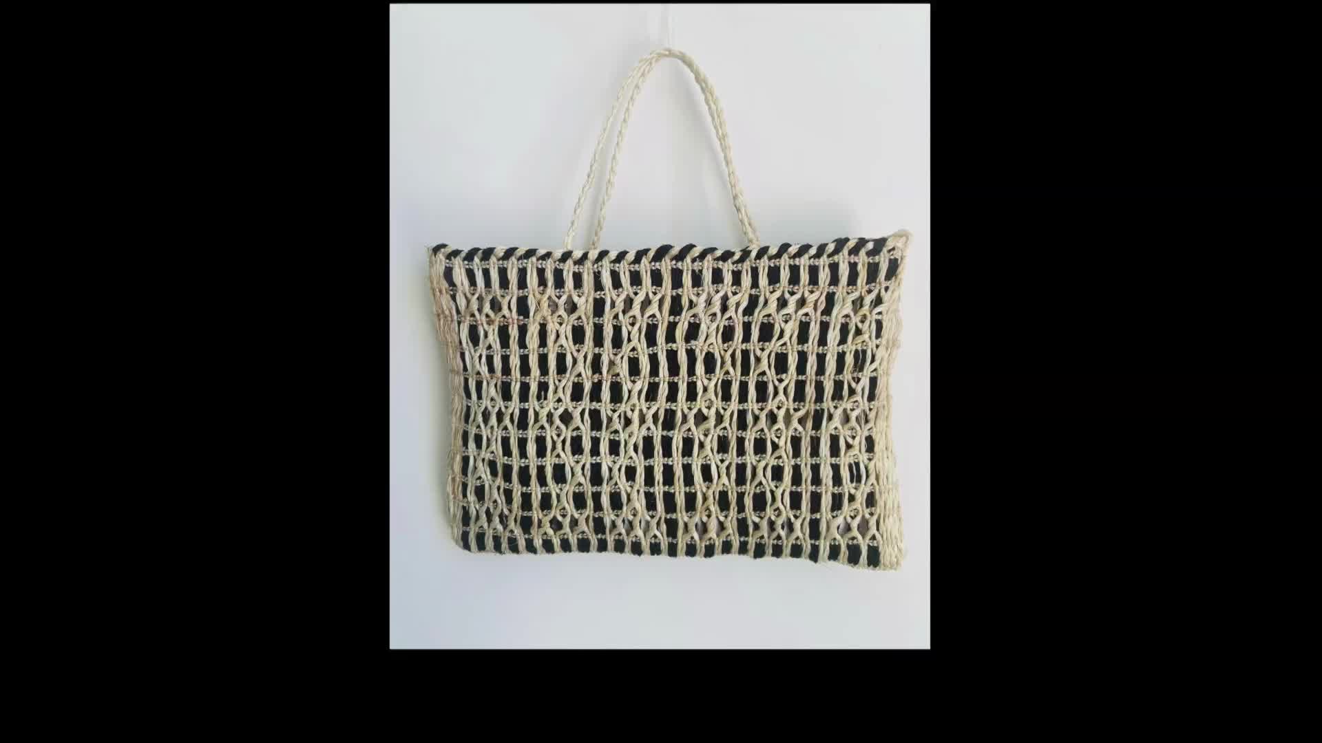 A woven tote bag, with a black interior, hangs against a white wall. The bag's handle is made of twisted rope.
