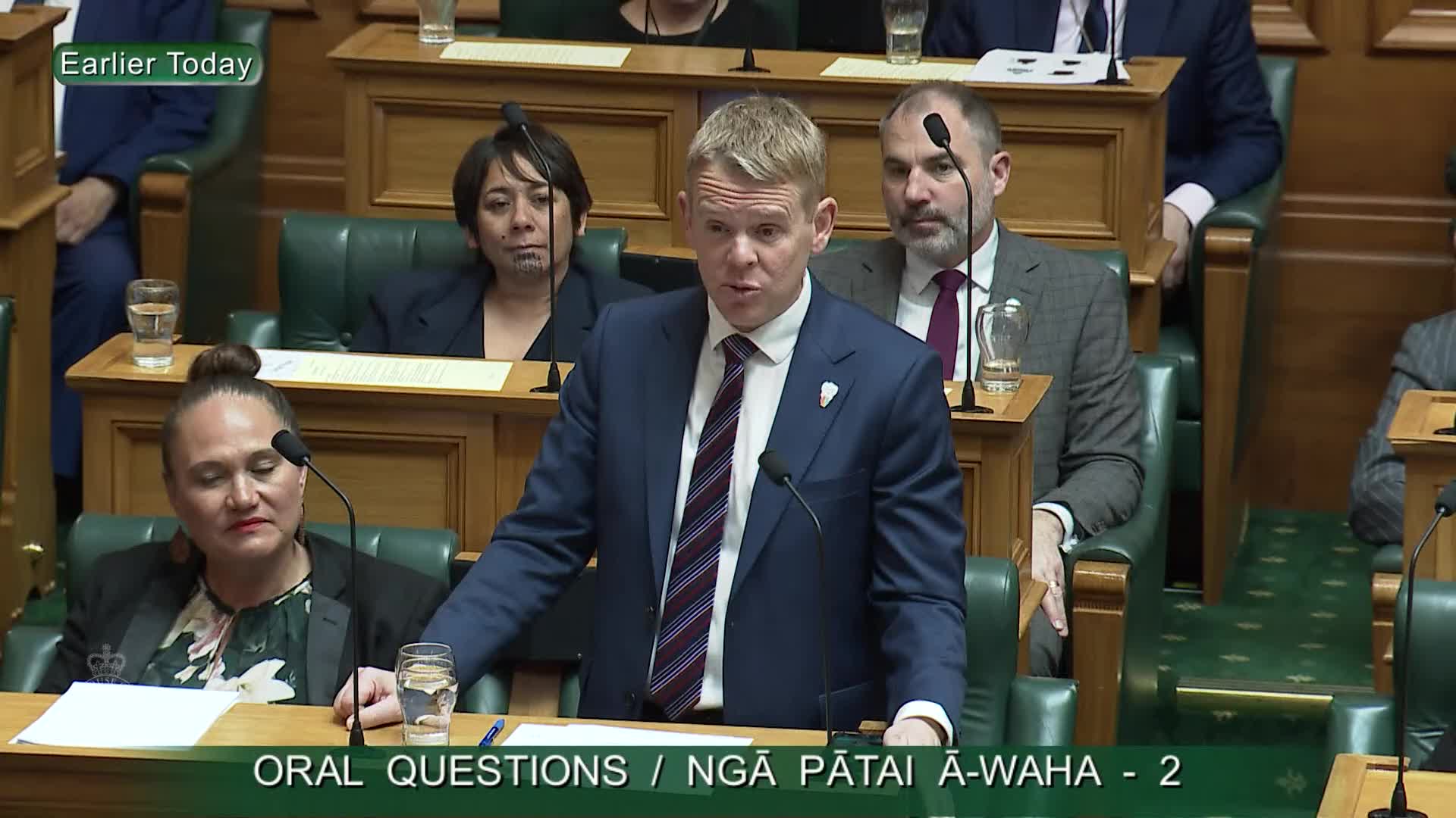 A man in a blue suit stands at a podium, speaking into a microphone. Other members of Parliament sit in the background, some looking on with varying expressions.
