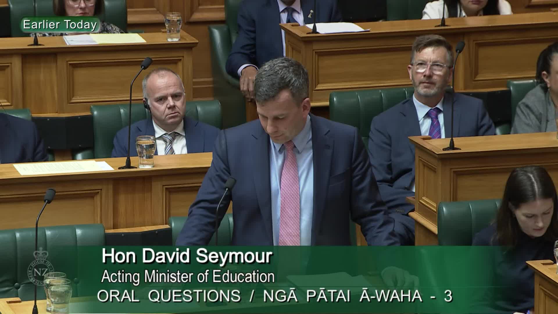 David Seymour, Acting Minister of Education, stands at the podium, speaking during Oral Questions. Other members of Parliament sit in the background, some looking on intently.
