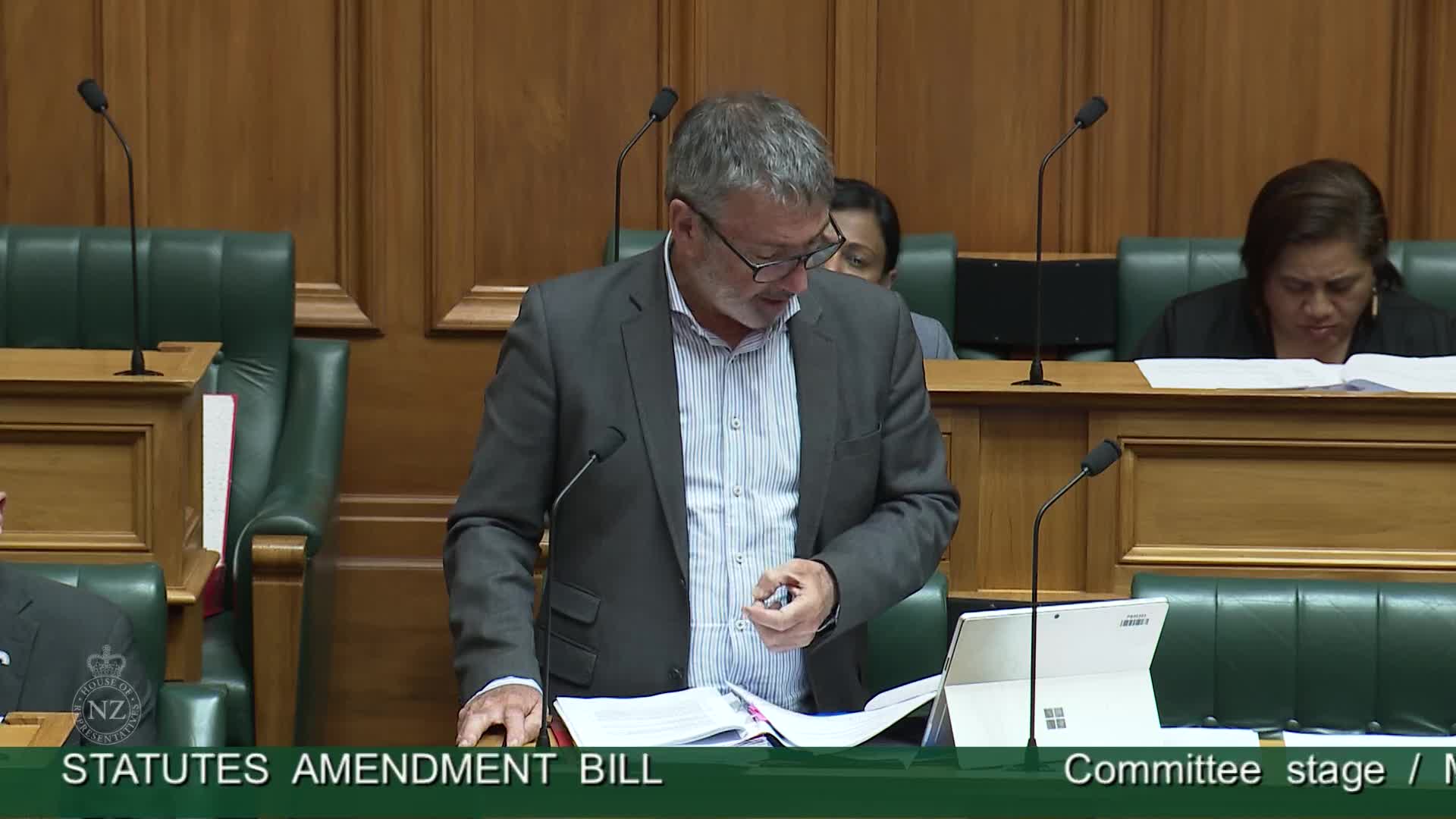 The man in the grey jacket is speaking, holding a pen and looking down at papers on the lectern. Two other people sit behind him, heads bowed, seemingly reading.
The man in the grey jacket is speaking, holding a pen and looking down at papers on the lectern. Two other people sit behind him, heads bowed, seemingly reading.