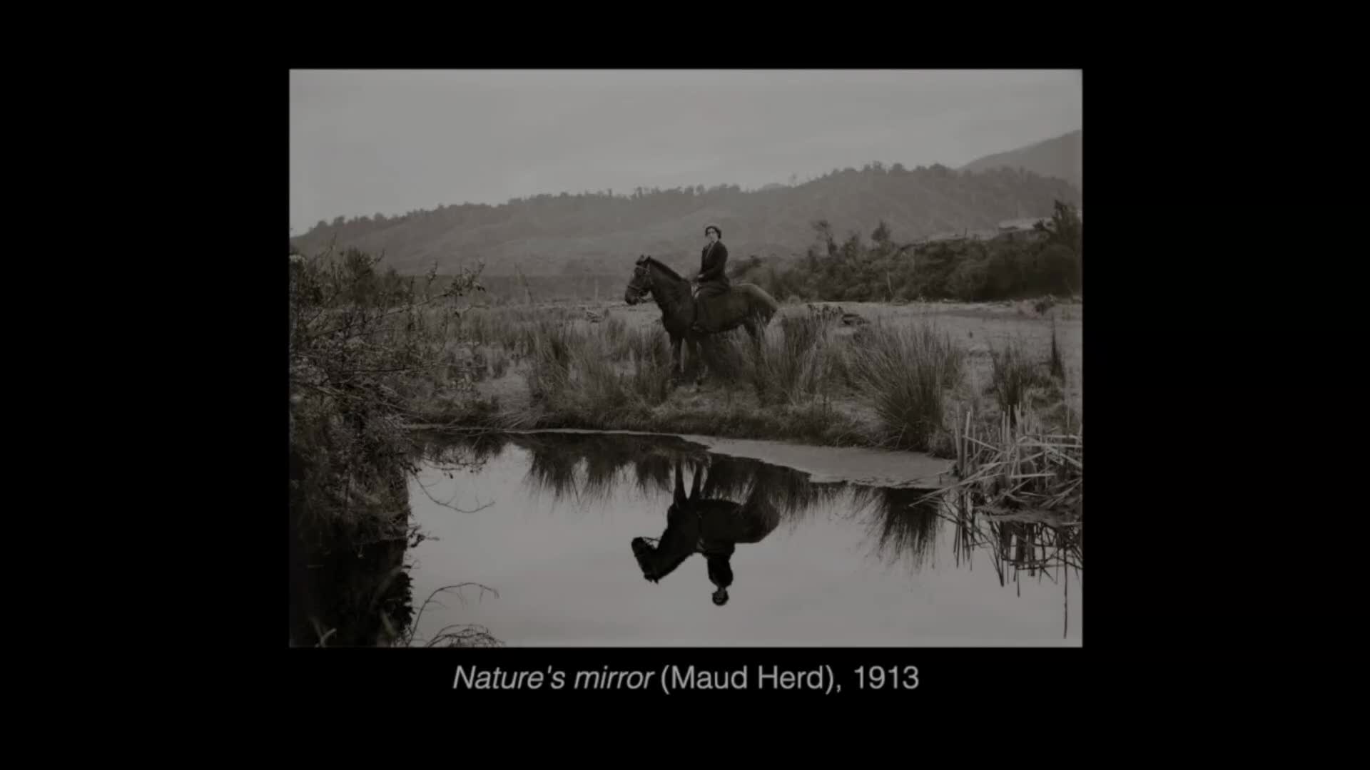A woman on horseback sits still, her reflection perfectly mirrored in the still water. The landscape around them is muted, with tall grasses and distant hills under a cloudy sky.
