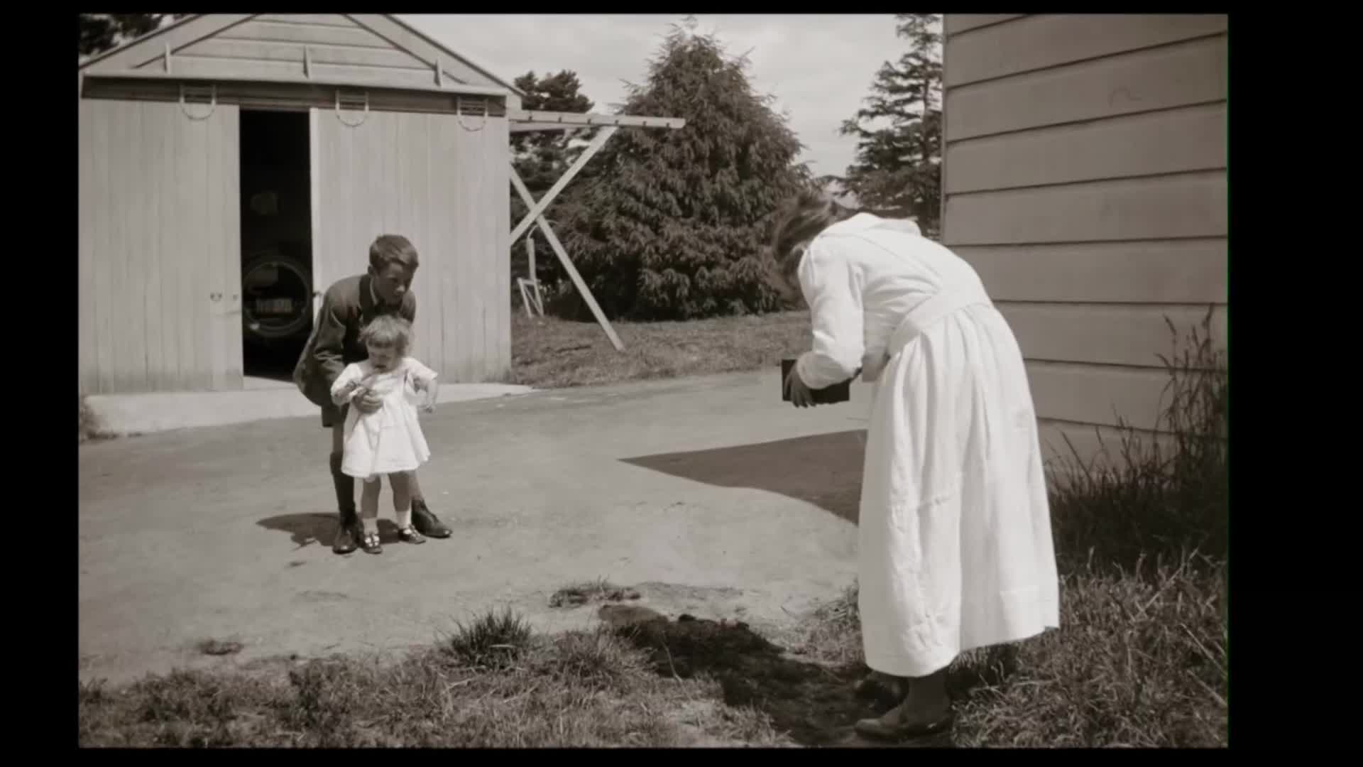 A young man holds a child in a white dress, posing for a photograph. A woman in a long white dress is focused on her camera, capturing the moment.