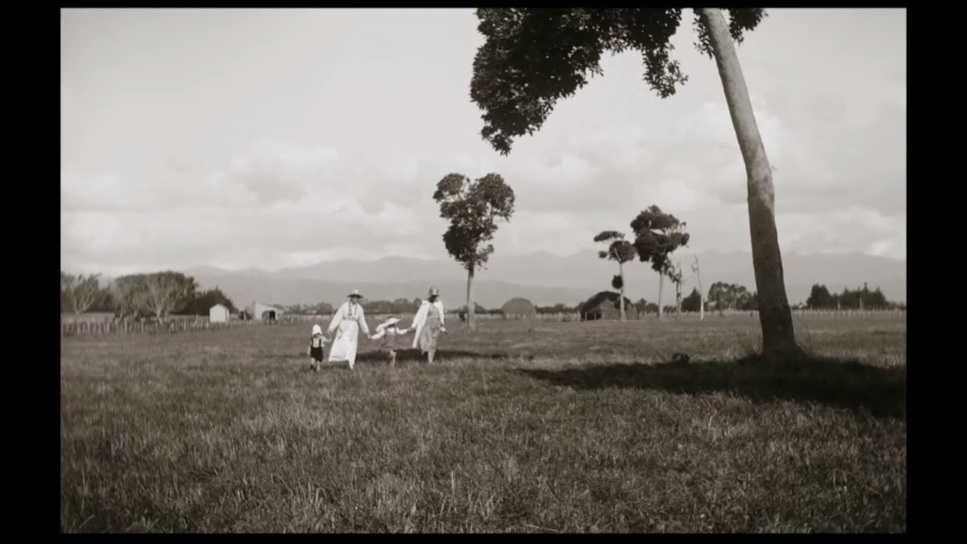 Two women and a child walk across a grassy field in New Zealand. The woman on the left holds the child's hand, while the woman on the right carries a kite.