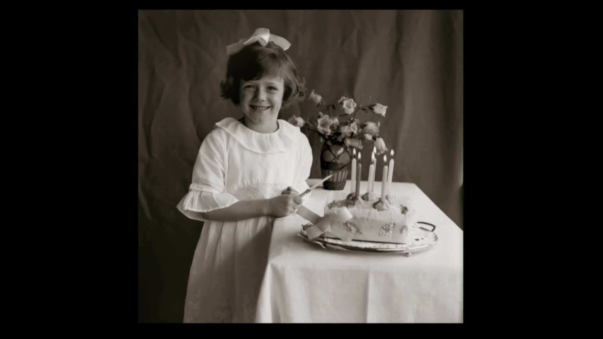 A young girl in a white dress with a pink bow smiles as she holds a knife over a cake. Seven lit candles flicker on the cake, which sits on a table covered with a white cloth.