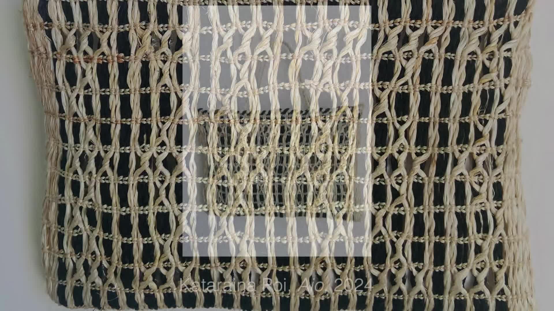 A woven panel of natural fibers hangs against a dark background. The intricate pattern of the weaving creates a textured surface, with a lighter, more open weave overlaying a denser grid.