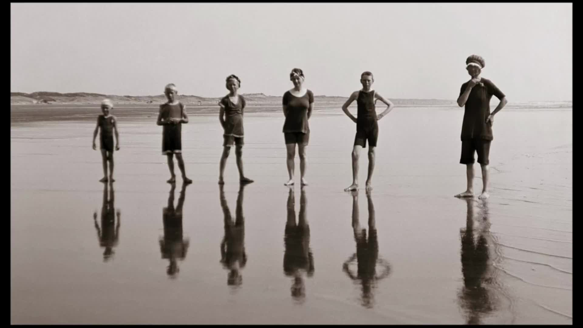 Seven children stand in a line on a wet, sandy beach, their reflections stretching out before them. The youngest, in a cap and shorts, is on the far left, while the tallest, wearing a cap and a dark swimsuit, stands on the right.